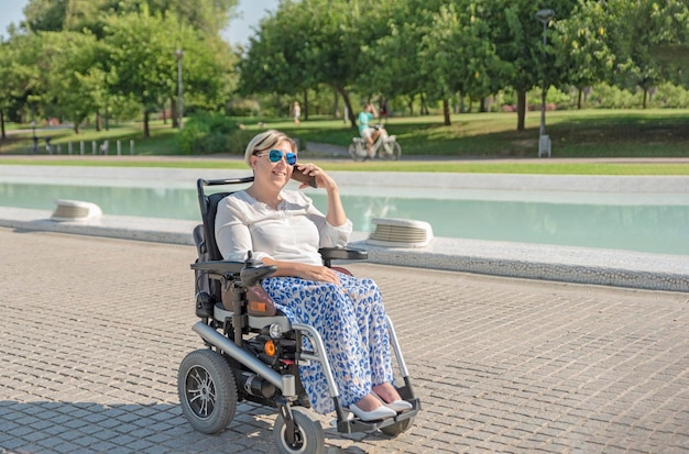 A smiling woman in an electric wheelchair with disability talks on the phone while enjoying a sunny day stroll in an urban park