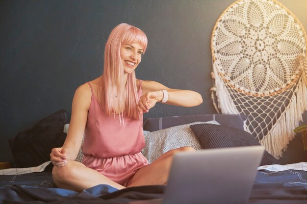 Улыбающаяся женщина в элегантной розовой пижаме проверяет время на умных часах в спальне