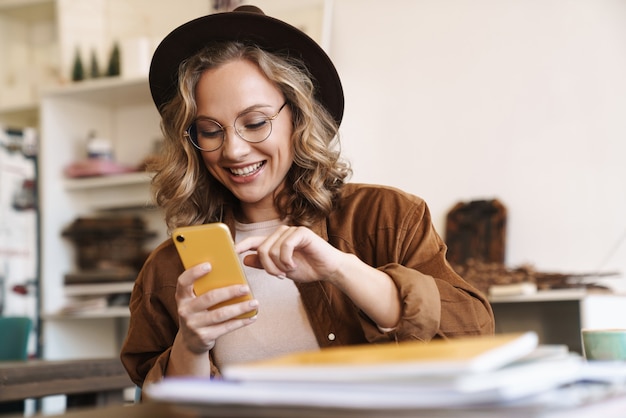 Photo smiling woman in eyeglasses and hat using cellphone while studying with exercise books at home