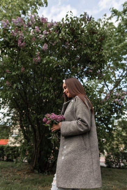 세련된 회색 코트를 입은 세련된 반다나를 입은 웃는 여성은 푸른 나무 근처에 라일락 꽃다발을 들고 있습니다. 패션 옷을 입고 달콤한 미소를 가진 행복한 귀여운 소녀는 공원에서 아름다운 꽃과 함께 산책합니다.
