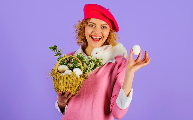 Donna sorridente in berretto alla moda con coniglietto di pasqua sulle spalle e uovo di pasqua in mano