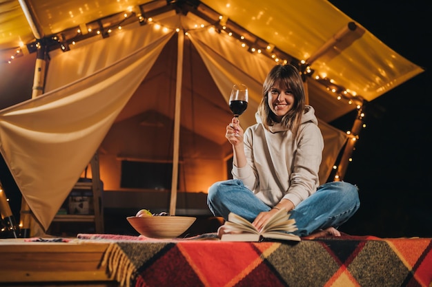 Smiling woman freelancer drinking wine and read book sitting in\
cozy glamping tent in autumn evening luxury camping tent for\
outdoor holiday and vacation lifestyle concept