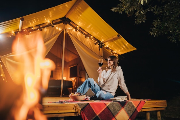 Smiling woman freelancer drinking wine and read book sitting in\
cozy glamping tent in autumn evening luxury camping tent for\
outdoor holiday and vacation lifestyle concept