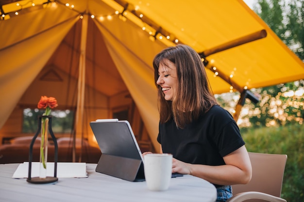 Smiling Woman freelancer using a laptop on a cozy glamping tent in a sunny day Luxury camping tent for outdoor summer holiday and vacation Lifestyle concept