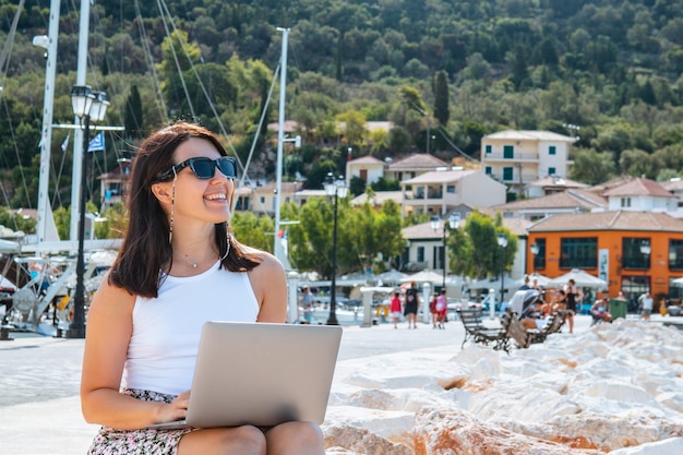 Smiling woman freelancer working on laptop outdoors at city harbor mountains and yacht on background