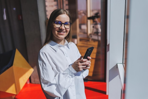 Smiling woman in glasses in casual wear watching a positive video on a phone connected to wifi a female blogger in a white shirt against a large window enjoying relaxation during remote work