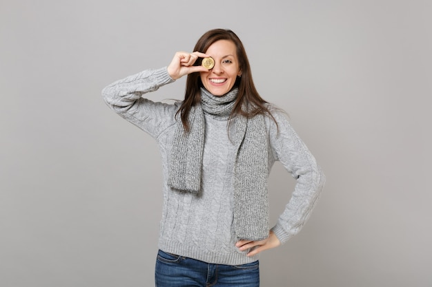 Smiling woman in gray sweater, scarf covering eye with bitcoin, future currency isolated on grey wall background. Healthy fashion lifestyle, people emotions, cold season concept. Mock up copy space.