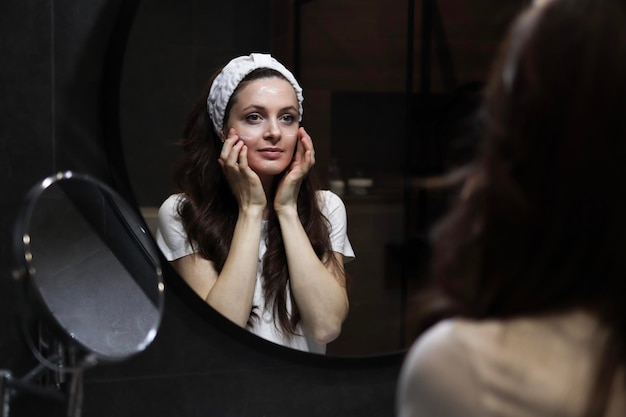 A smiling woman in a headband is taking care of her skin by doing face massage in front of a mirror in a loft interior bathroom Natural skincare Beauty home spa with vegan cosmetics