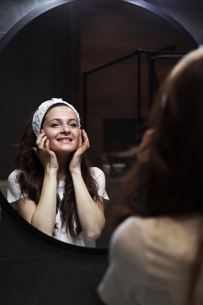 A smiling woman in a headband is taking care of her skin by doing face massage in front of a mirror in a loft interior bathroom Natural skincare Beauty home spa with vegan cosmetics