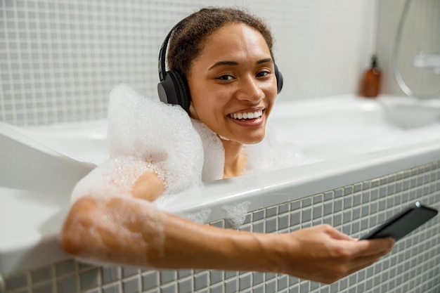 헤드폰을 끼고 웃고 있는 여성은 홈 스파와 뷰티 루틴에서 목욕을 할 때 스마트폰을 사용하고 있습니다.