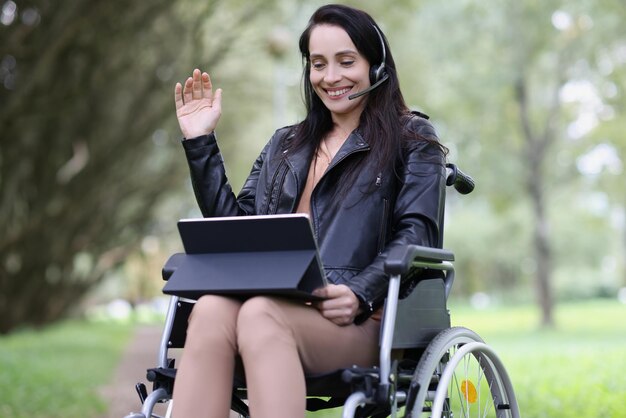 Улыбающаяся женщина в наушниках и ноутбуке в инвалидной коляске в парке снимает общение и работает на