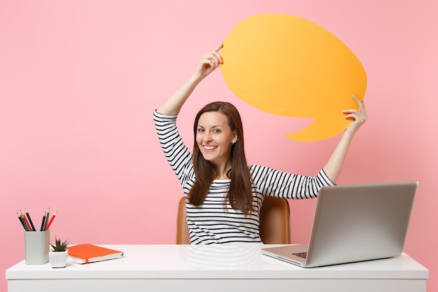 웃고 있는 여성은 파스텔 핑크색 배경에 격리된 PC 노트북이 있는 흰색 책상에서 노란색 빈 세이 클라우드 말풍선을 들고 있습니다. 성취 비즈니스 경력 개념입니다. 광고 공간을 복사합니다.