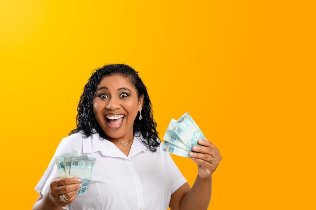 Smiling woman holding 100 reais notes in her hands Brazilian money isolated on orange background