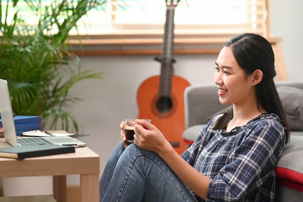 Smiling woman holding coffee cup and using laptop while sitting in living room.