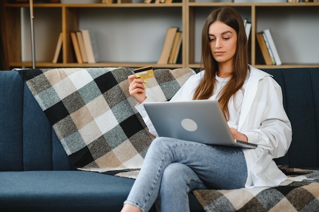 Smiling woman holding credit card and using laptop on sofa in living room