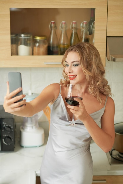 Donna sorridente che tiene un bicchiere di vino rosso e si fa selfie per i social media