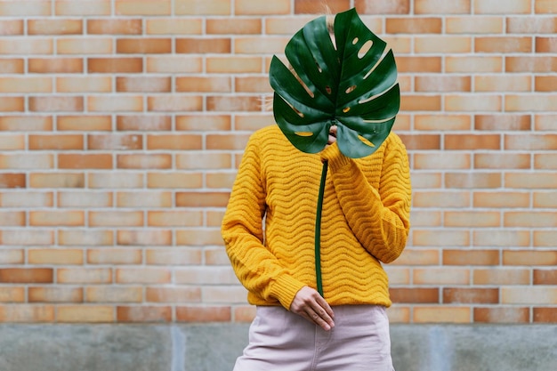 Smiling woman holding monstera leaf in front of brick wall Concept of Ecology Sustainability