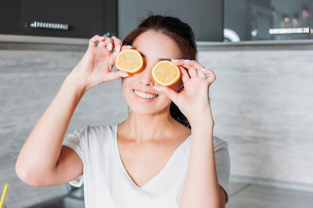 Photo smiling woman holding orange slices against eyes at home