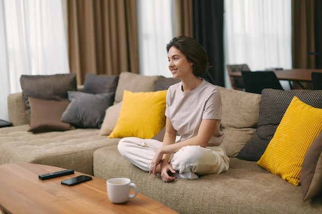 Smiling woman homeowner tenant sitting on sofa in cozy living room watching television resting on