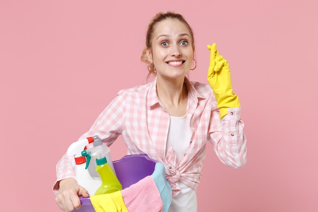 Smiling woman housewife in rubber gloves hold basin with detergent bottles washing cleansers doing housework isolated on pink background. Housekeeping concept. Making wish, keeping fingers crossed.