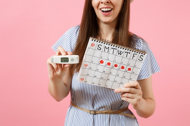 Фото Улыбающаяся женщина в платье держит в руке термометр, женский календарь периодов для проверки дней менструации, изолированных на розовом фоне. медицинское здравоохранение, гинекологическая концепция овуляции. скопируйте пространство.