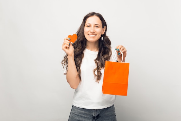 Smiling woman is holding orange shopping bag and heart over white background.