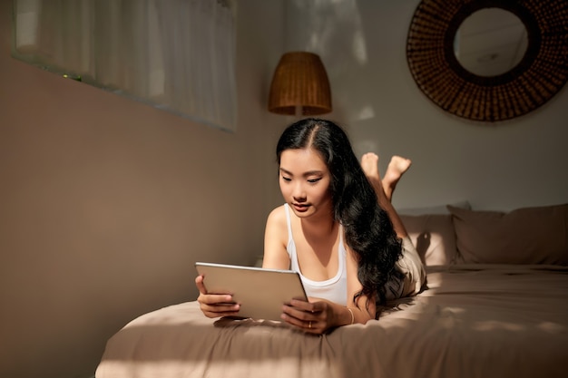 A smiling woman lies on the bed scrolling through her tablet.