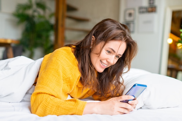 Smiling woman lies in bed with cell phone she woke up and was checking social media posts