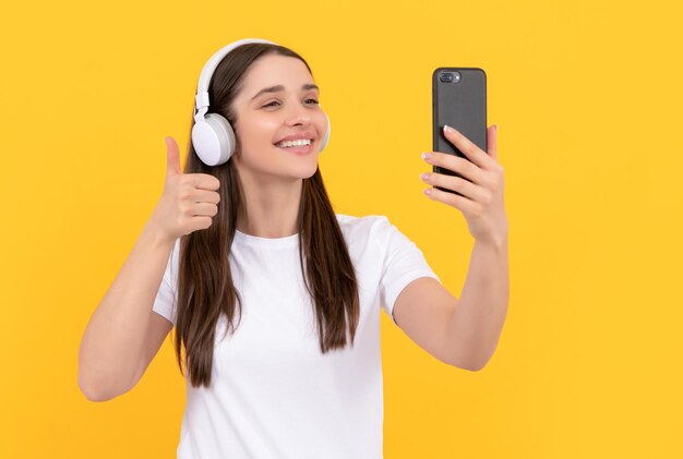 Smiling woman listen music in earphones with smartphone show thumb up on yellow background selfie