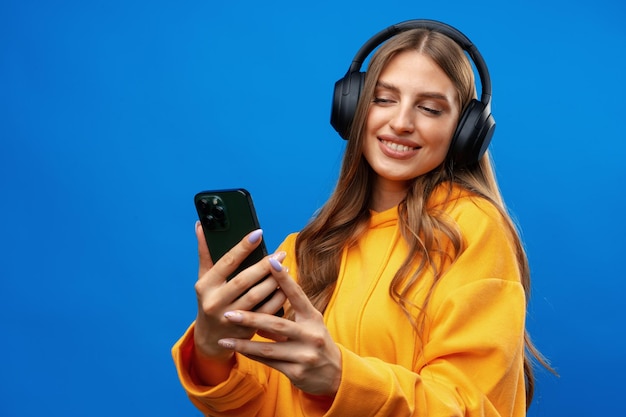 笑顔の女性がヘッドフォンで音楽を聴いて青い背景の上でスマートフォンを使用しています