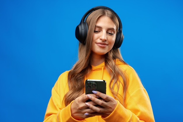 笑顔の女性がヘッドフォンで音楽を聴いて青い背景の上でスマートフォンを使用しています