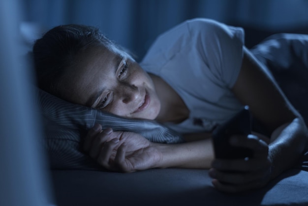 Photo smiling woman lying in bed at night she can't sleep and she is connecting online with her smartphone