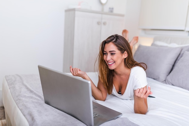 Smiling woman lying down the bed in front of her laptop with her legs raised. Happy casual beautiful woman working on a laptop sitting on the bed in the house. Freelance working from home concept