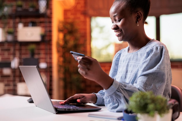 Smiling woman making purchase in internet shop using laptop. Young african american girl holding debit card and searching goods in online store, electronic shopping concept