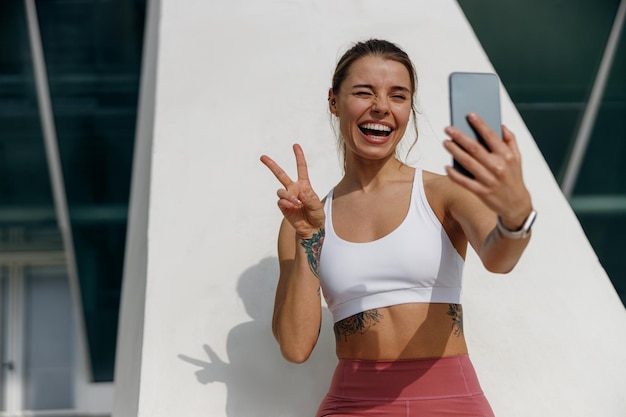 smiling woman making selfie on a smart phone after morning run outdoors and showing sign peace
