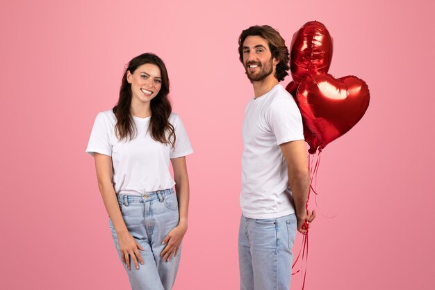 Smiling woman and man in white tshirts with the man holding a bunch of red heartshaped balloons