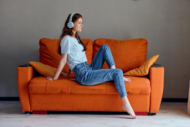 Улыбающаяся женщина на оранжевом диване слушает музыку в наушниках