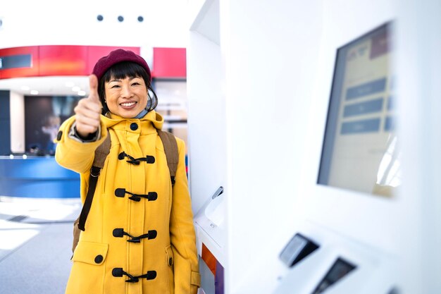 Smiling woman passenger with backpack standing by the airport boarding pass machine