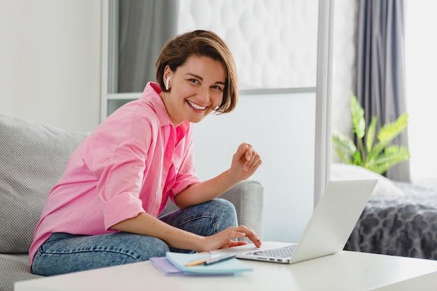 Donna sorridente in camicia rosa seduto rilassato sul divano a casa a tavola lavorando online su laptop da casa