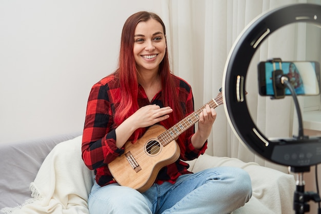 Smiling woman playing ukulele recording on phone camera the musician performs a live concert from home