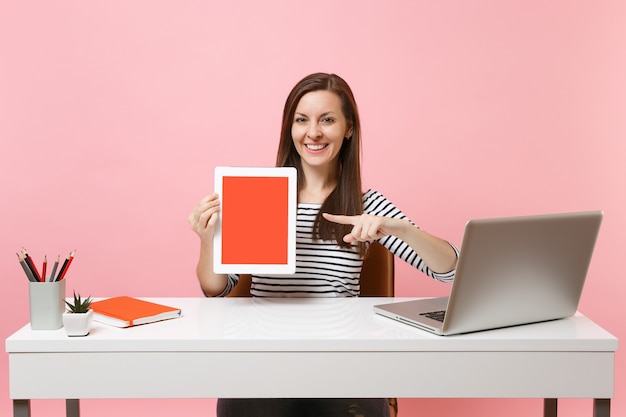Smiling woman pointing index finger on tablet computer with blank empty screen, sit work at white desk with contemporary pc laptop 