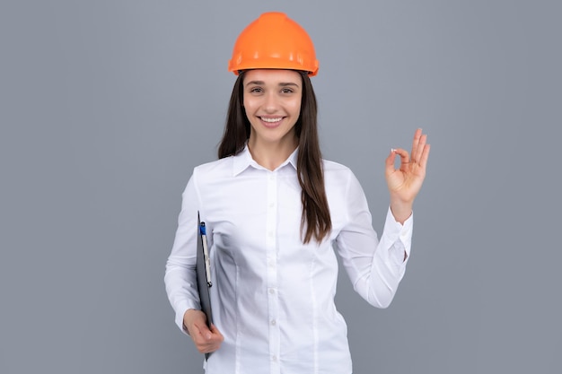 Smiling woman in protective helmet with clipboard isolated on grey background Young woman