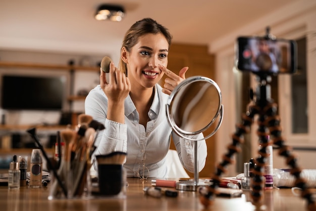 Smiling woman recording makeup vlog while applying face powder at home