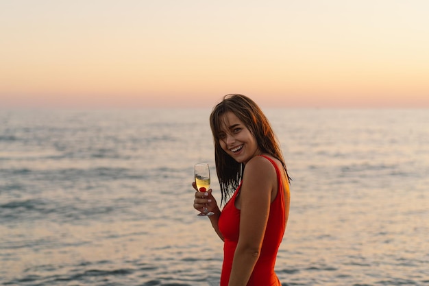 Smiling woman in red swimsuit holding wine glass by the sea at sunset