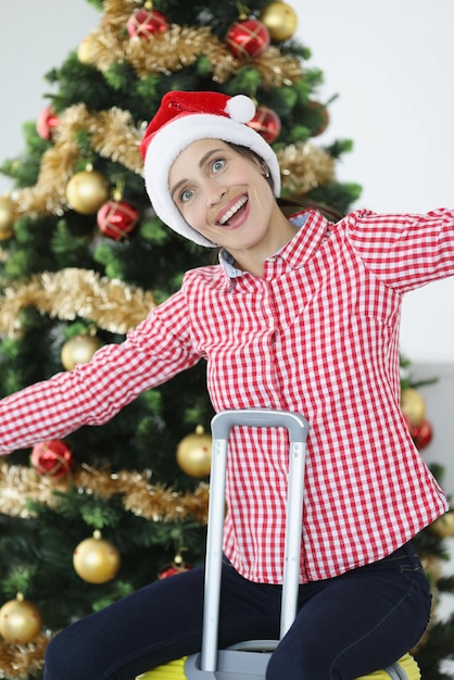 Smiling woman in santa claus hat sits on suitcase against background of christmas tree trips