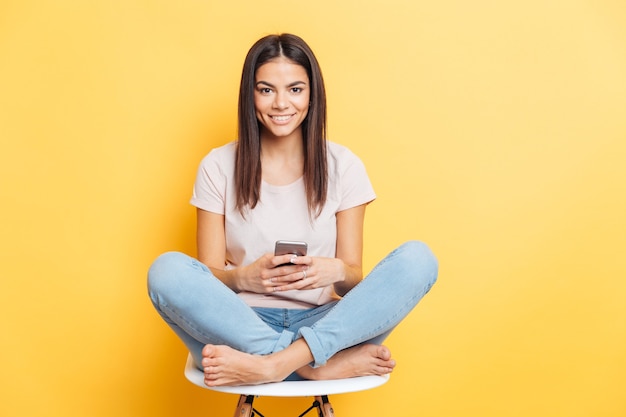 Smiling woman sitting on the chair and using smartphone over yellow wall