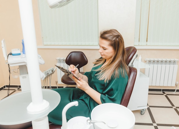 Donna sorridente che si siede sulla sedia del dentista e che compila il modulo di domanda medica