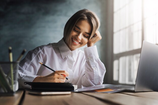 Smiling woman sitting at desk, making notes with pen in copybook