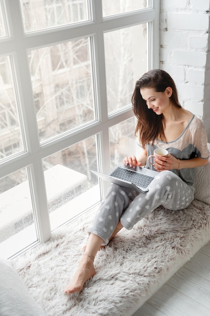 Smiling woman sitting near white window and chatting on laptop.