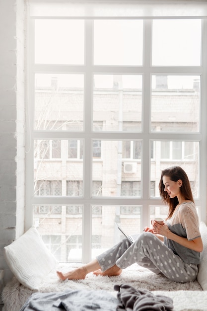 Smiling woman sitting near white window and chatting on laptop.